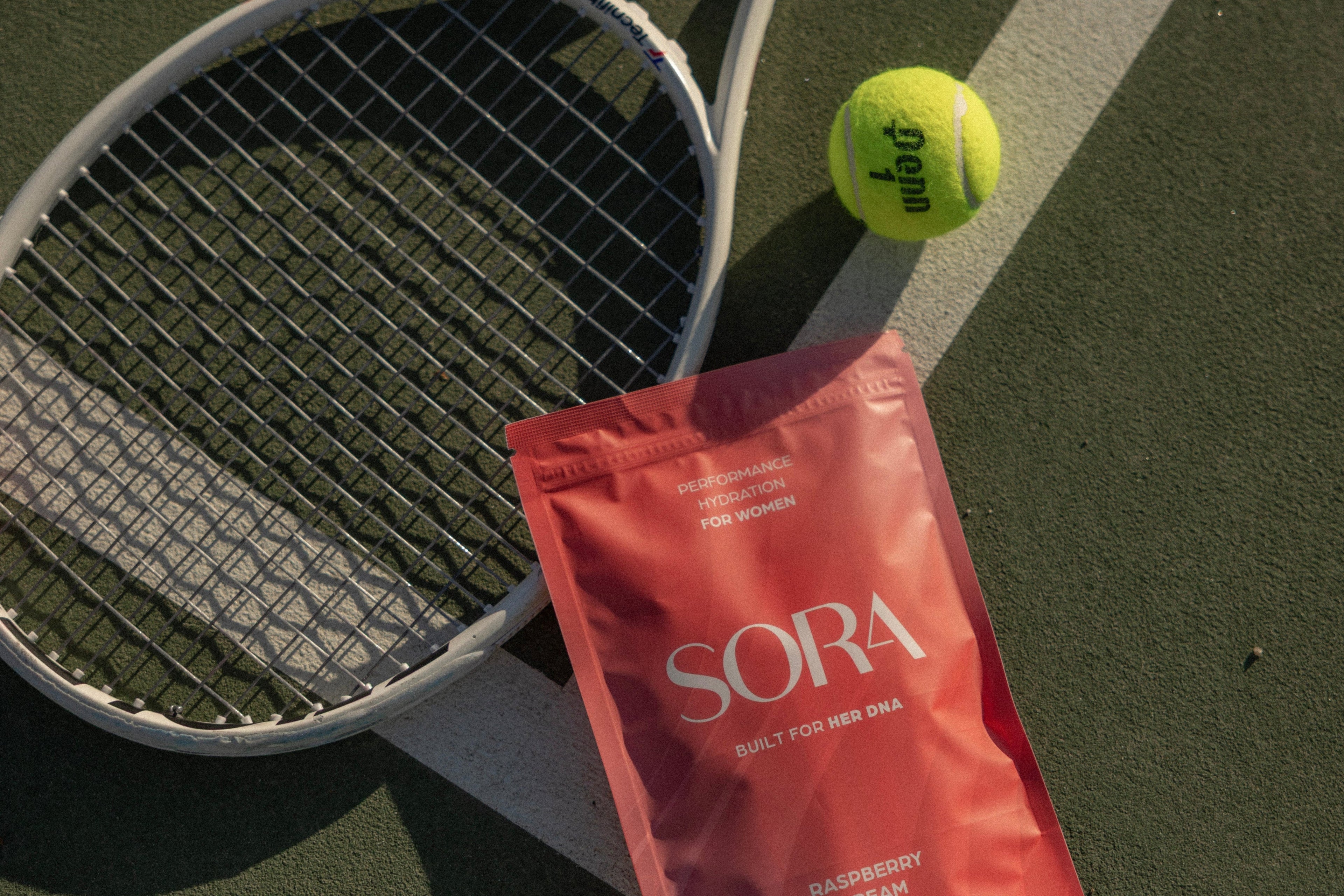 A red SORA protein powder pouch lies on a tennis court next to a tennis racket and a yellow tennis ball.
