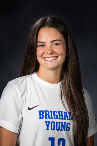 A smiling young woman with long dark brown hair wearing a white athletic jersey with blue 'BRIGHAM YOUNG' lettering and a black Nike swoosh, photographed against a dark studio background.