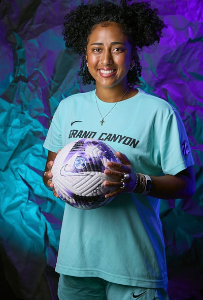 A smiling woman with curly hair stands holding a soccer ball, wearing a light aqua Nike Grand Canyon jersey and matching shorts against a purple and blue textured backdrop.