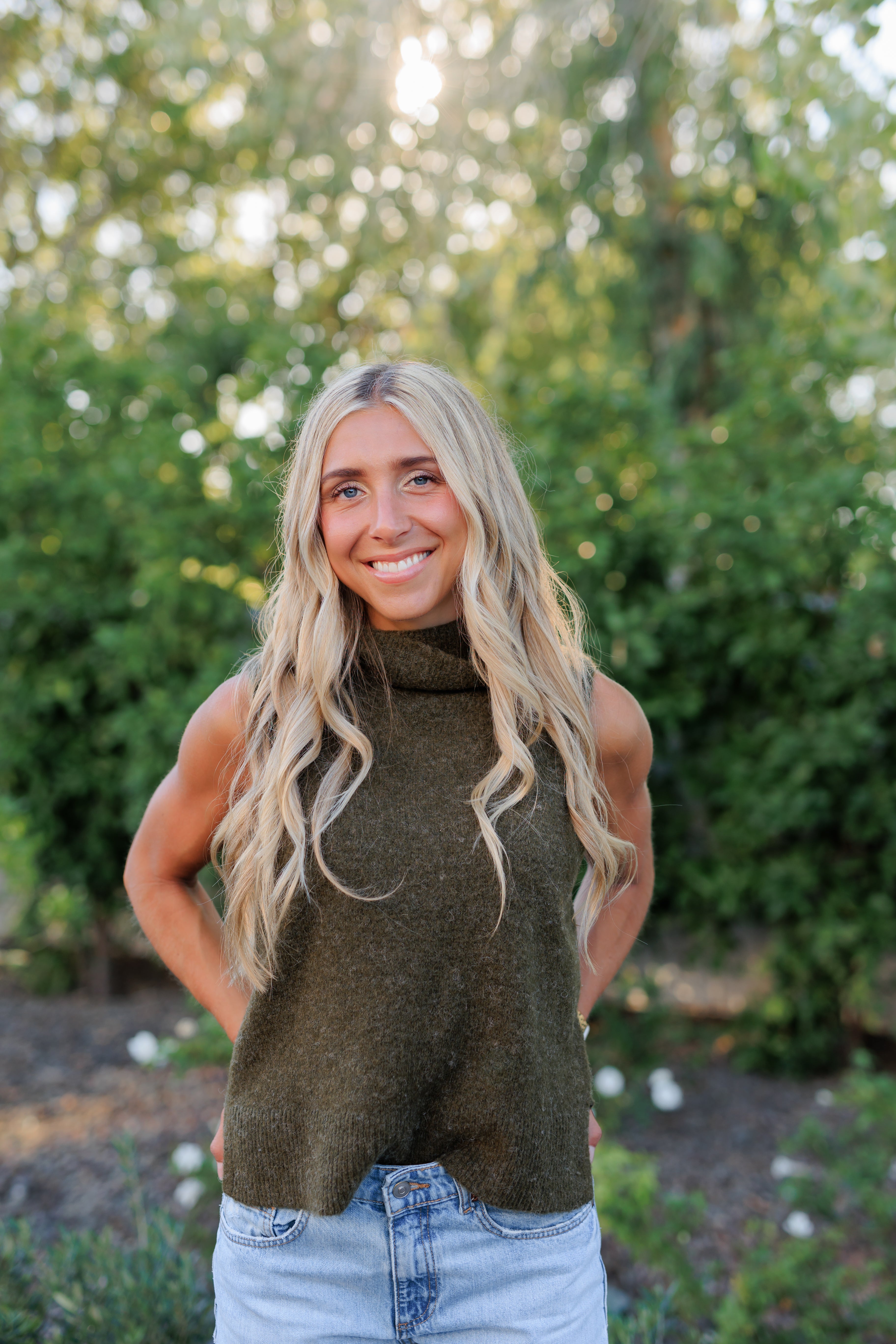 Woman wearing a green turtleneck sweater and blue jeans standing outdoors with trees in the background